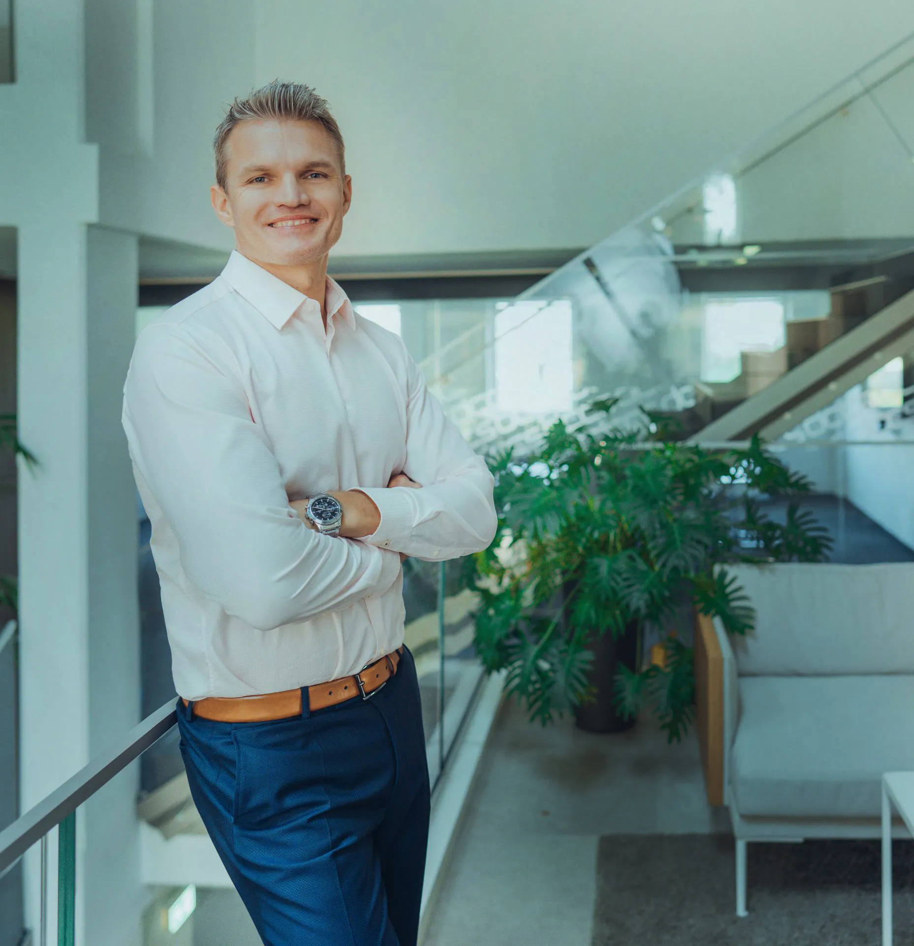 Lächelnder Mann mit weißem Hemd, blauer Hose und braunem Gürtel lehnt im modernen Büro an einem Geländer.