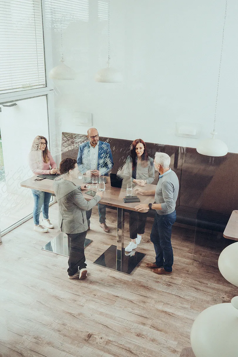 Fünf Personen führen ein Stehteam-Meeting in einem hellen Raum mit großem Fenster und Holzboden.