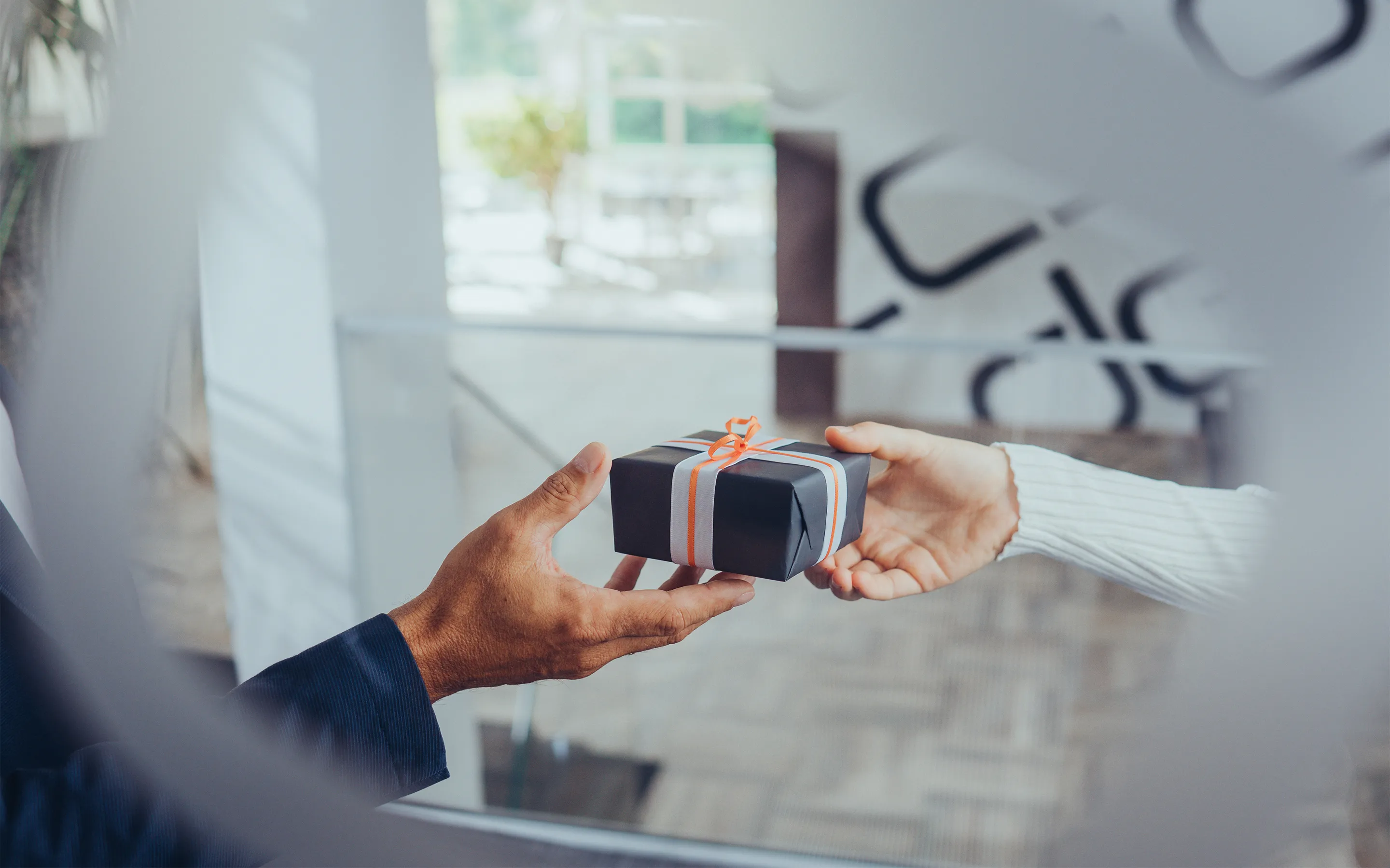 Zwei Personen reichen sich ein schwarz verpacktes Geschenk mit weißem und orangem Band.