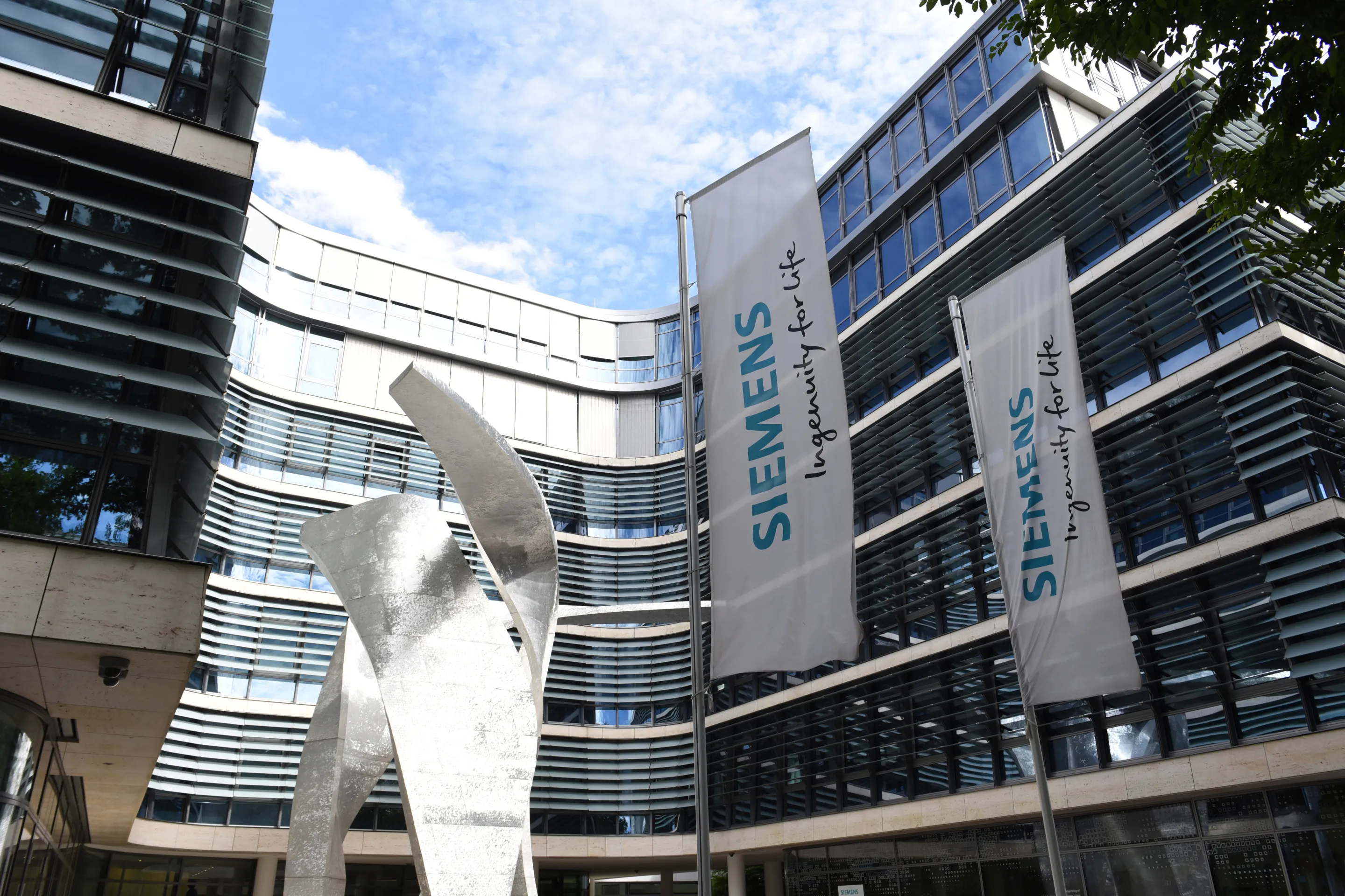 Zwei Siemens-Fahnen vor einem modernen Bürogebäude mit einem großen, silbernen abstrakten Metallkunstwerk im Innenhof.