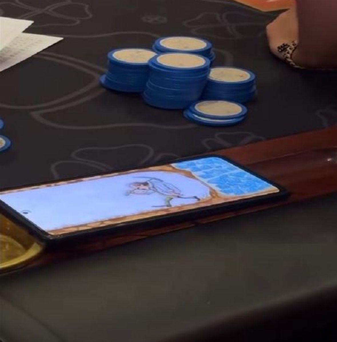 Person wearing headphones writing in a notebook at a desk with a laptop and a smartphone connected by a charging cable.