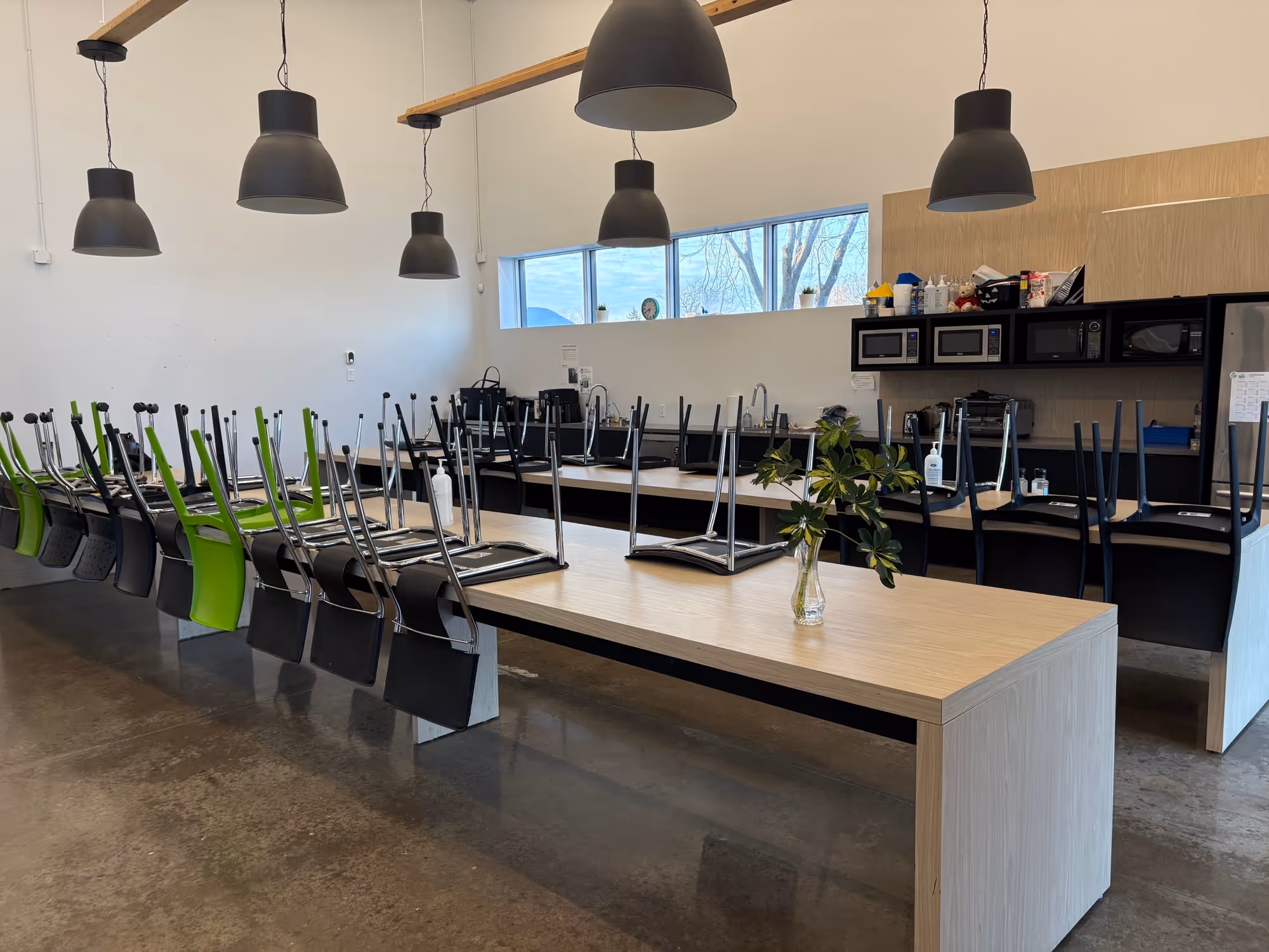 Salle commune avec tables en bois clair, chaises retournées sur les tables, luminaires suspendus noirs et une fenêtre longitudinale laissant entrer la lumière naturelle.