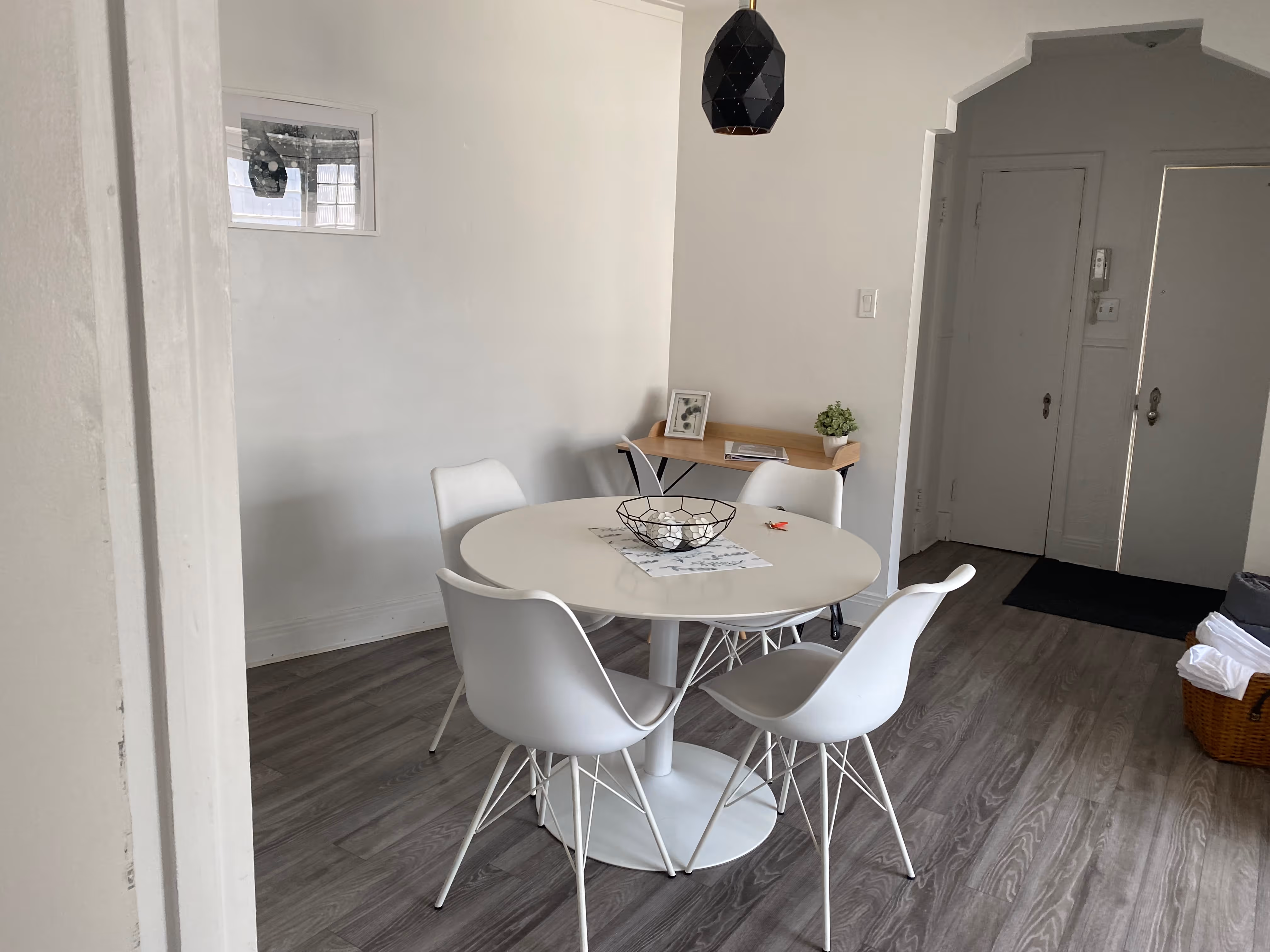 Salle à manger moderne avec table ronde blanche entourée de quatre chaises blanches, une petite table en bois avec cadre photo et plante en pot, sol en bois gris et panier avec serviettes à côté d'une porte d'entrée.