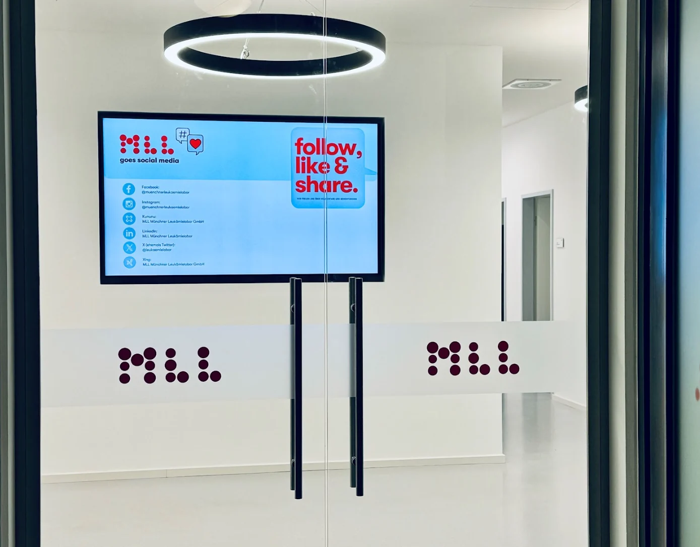 Display hinter einer Glastür im Münchner Leukämielabor zeigt Social-Media-Hinweise mit dem MLL Logo