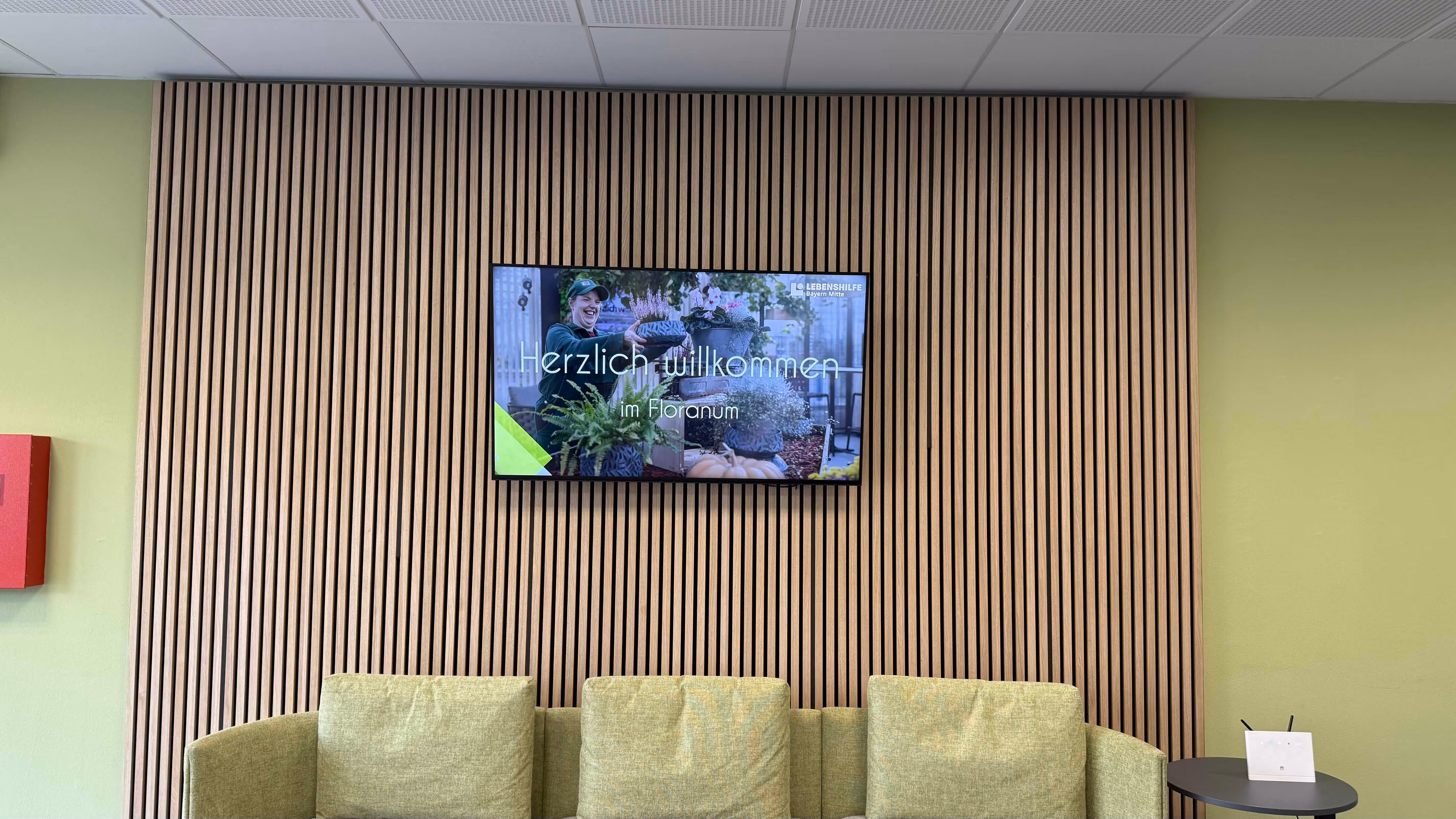 Welcome Screen über grünen Sesseln an einer Holzlamellenwand bei der Lebenshilfe Bayern Mitte