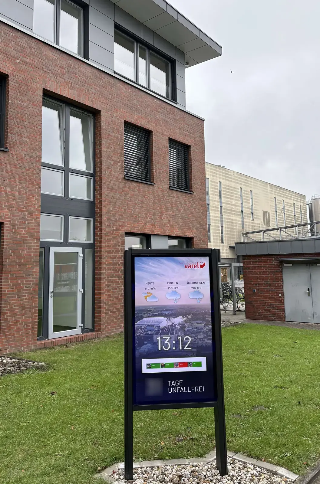 Freistehendes Digital Signage Display vor dem Backsteingebäude in Varel mit Wetter, Uhrzeit und unkenntlich gemachtem Unfallzähler
