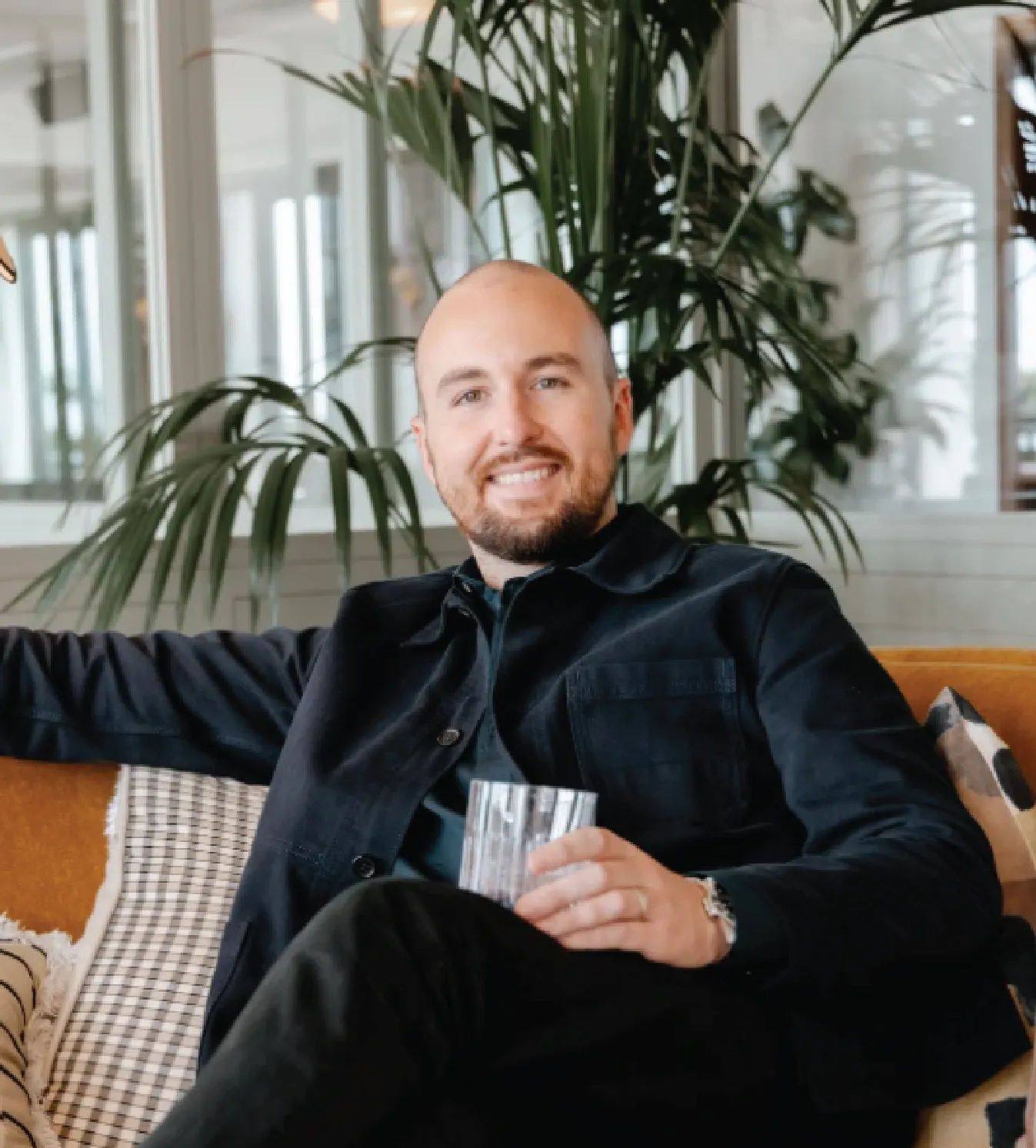 Smiling bald man with beard sitting on a couch holding a glass, with green plants and windows in the background.