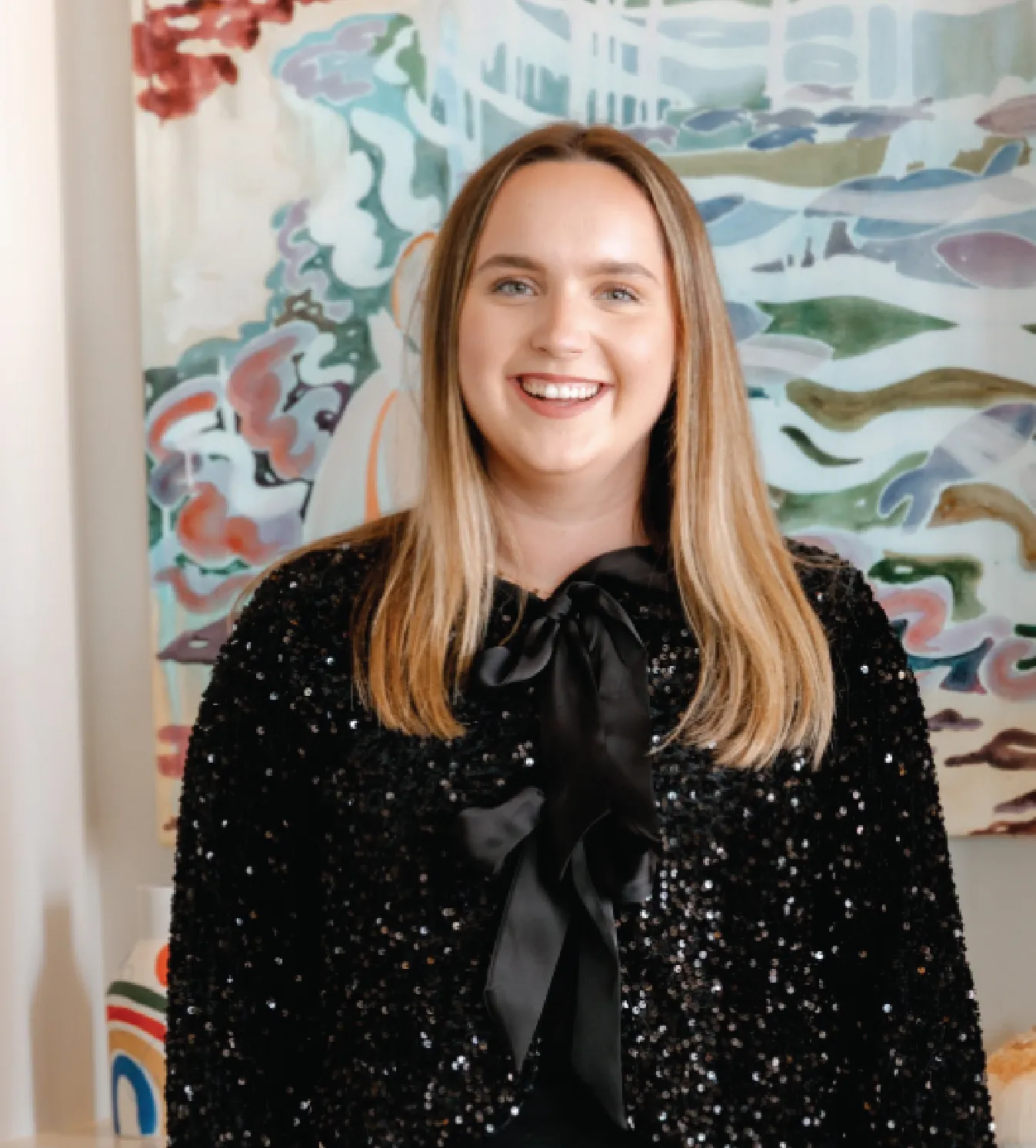 Smiling woman with long blonde hair wearing a black sequined top with a satin bow, standing in front of a colorful abstract painting.