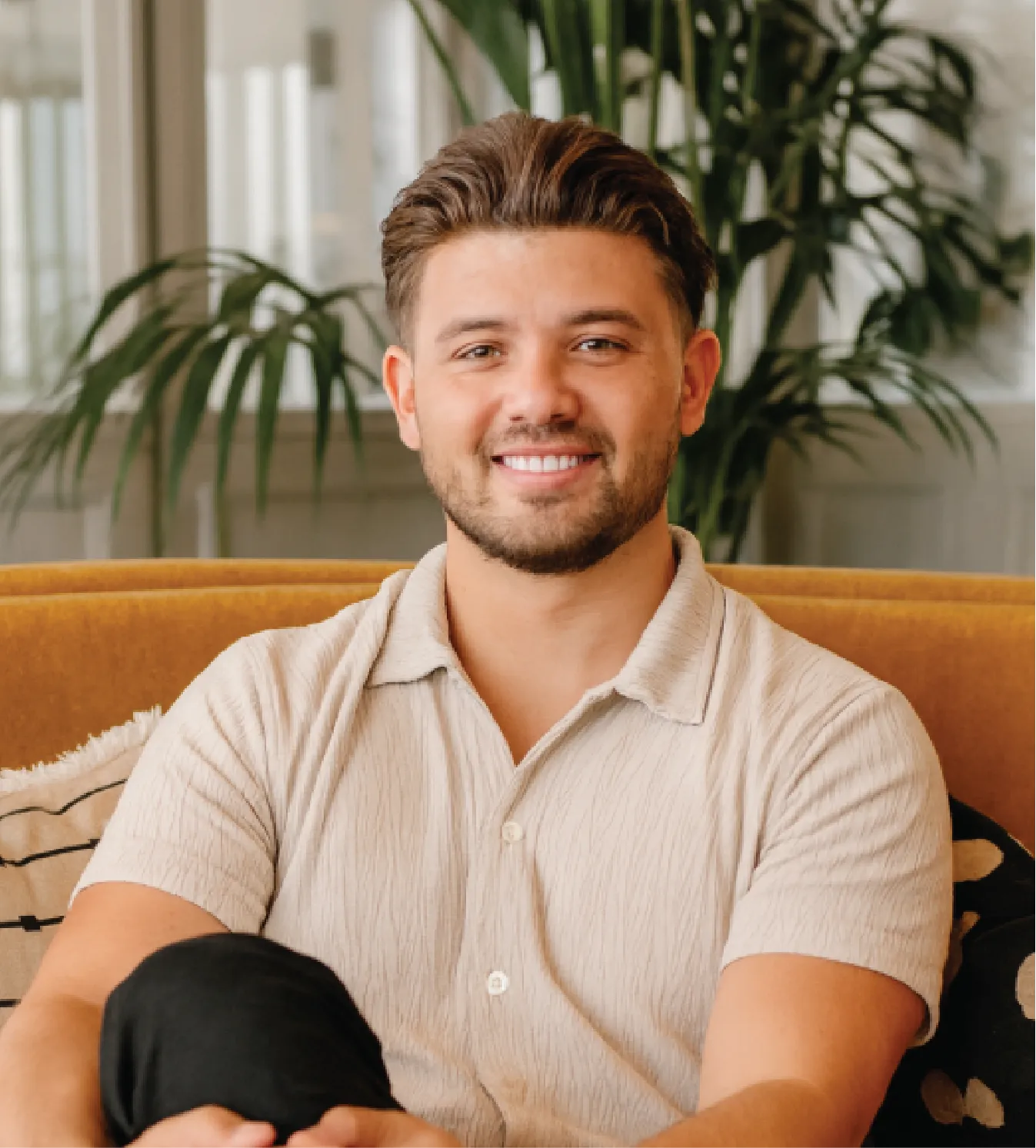 Smiling man with short brown hair and beard sitting on a mustard sofa with green plants in the background.