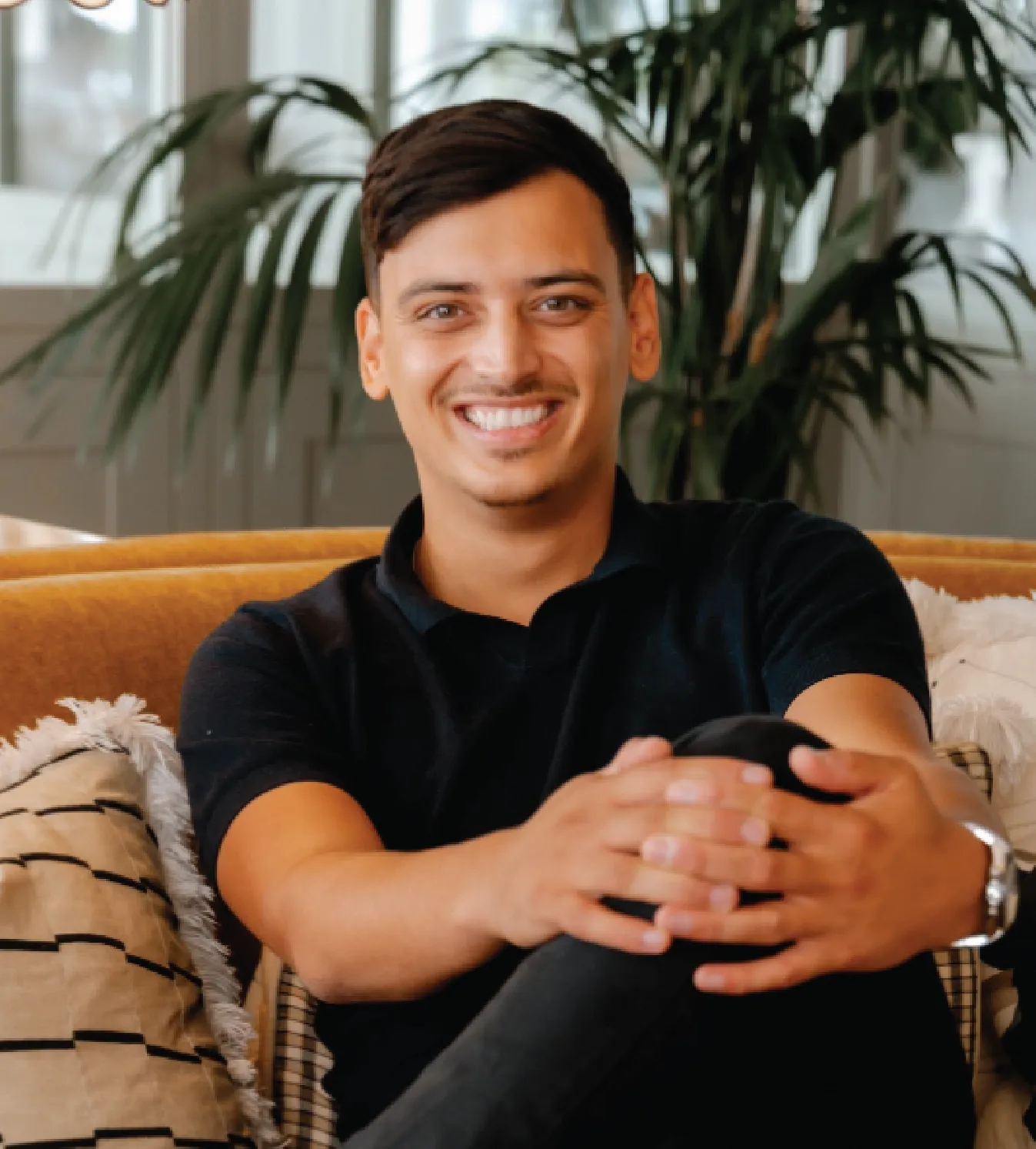 Smiling young man sitting cross-legged on a mustard yellow couch with patterned cushions and plants in the background.