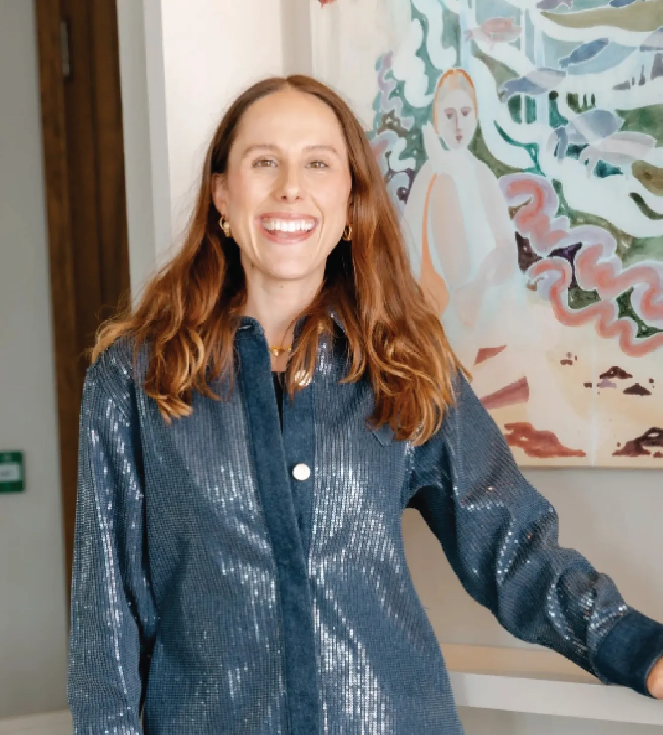 Smiling woman with long brown hair wearing a shiny blue shirt standing indoors in front of a colorful abstract painting.