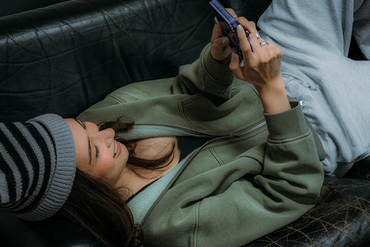 Smiling woman wearing a striped beanie and green jacket lying on a black couch while using a smartphone.