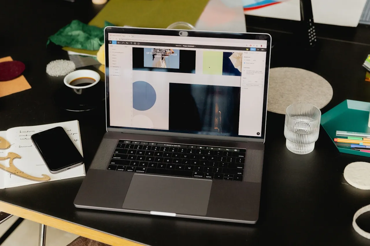 Open laptop on a black table displaying a design software interface, surrounded by a smartphone, notebook, coffee cup, glass of water, and colored pencils.