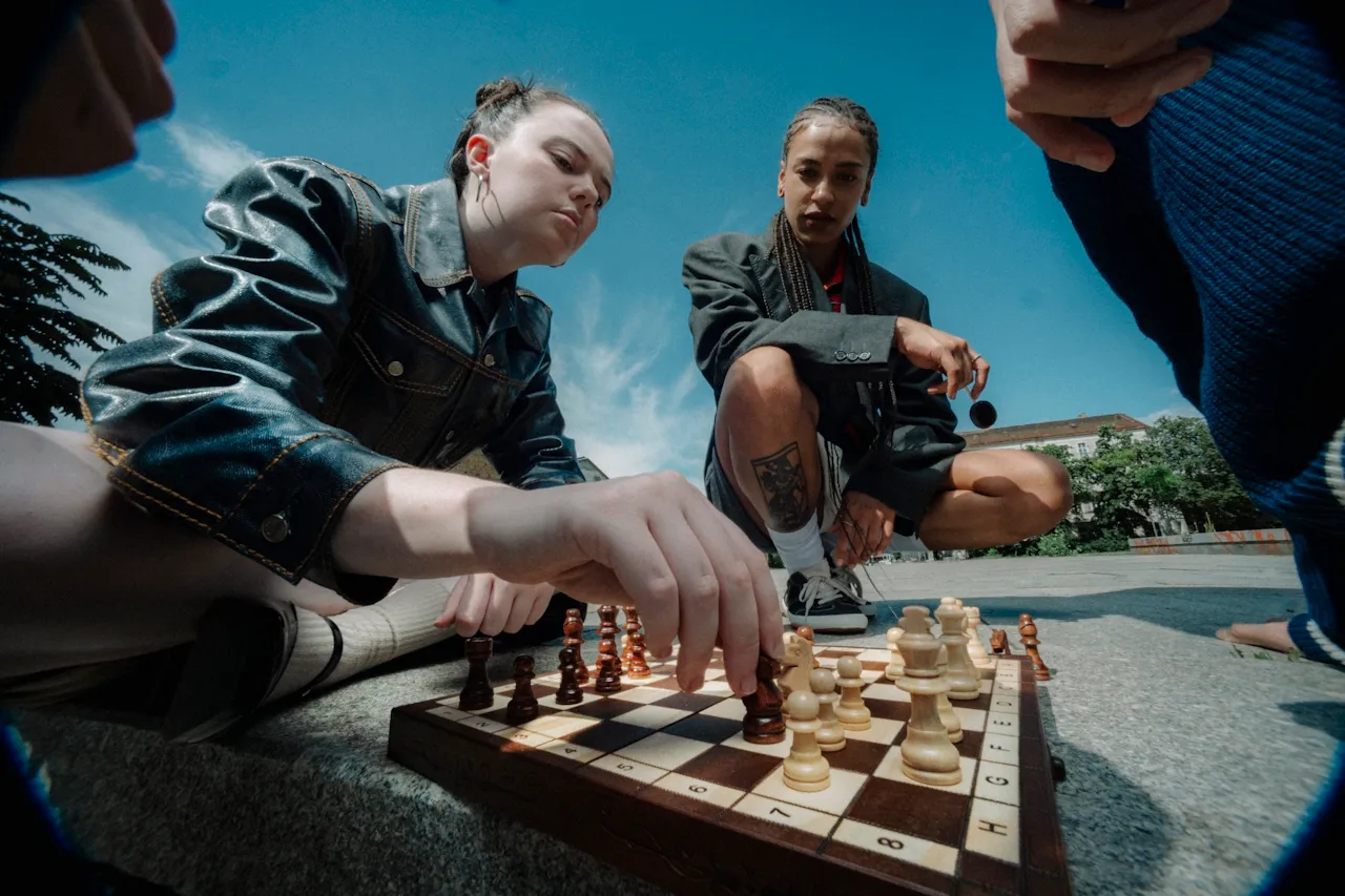 Two young women playing chess outdoors on a stone surface under a blue sky.