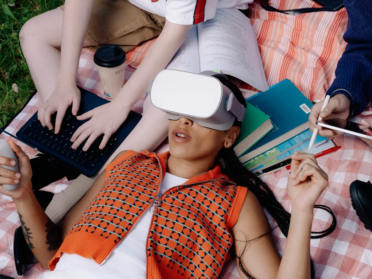 Person lying on a checkered blanket wearing a VR headset, surrounded by study materials and people using a tablet and keyboard.