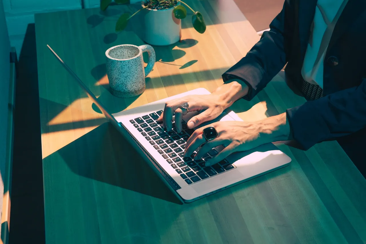 Person wearing a dark blazer typing on a laptop keyboard at a wooden desk with a speckled mug and a small plant nearby.