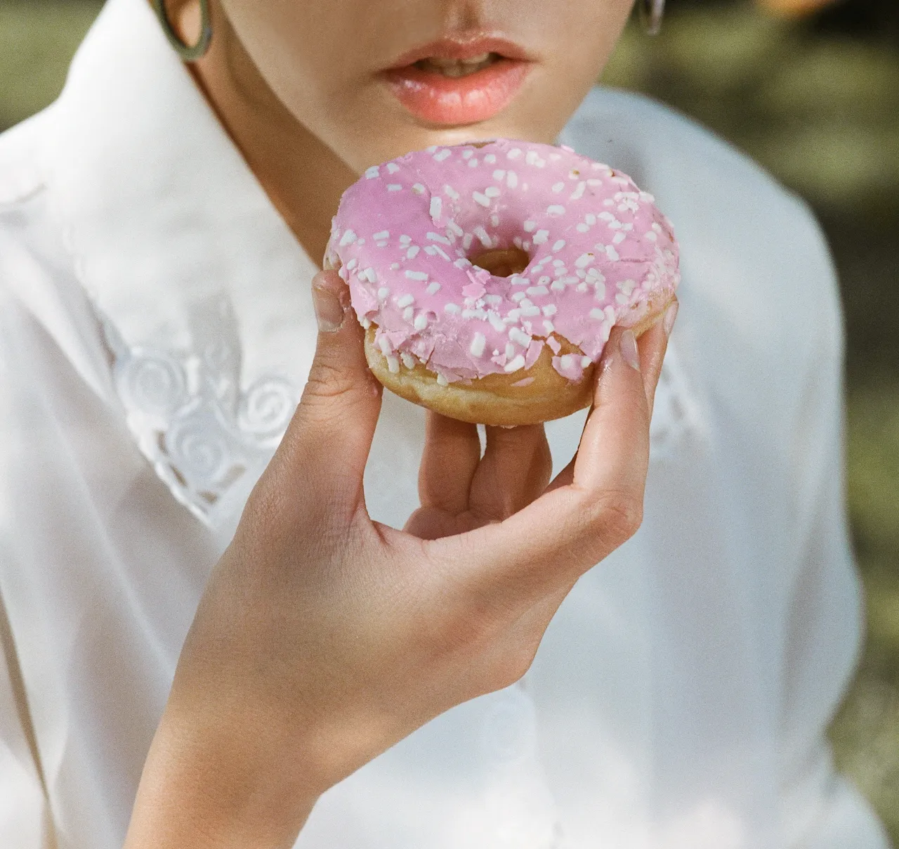 Person holding a pink-frosted donut with white sprinkles near their lips.