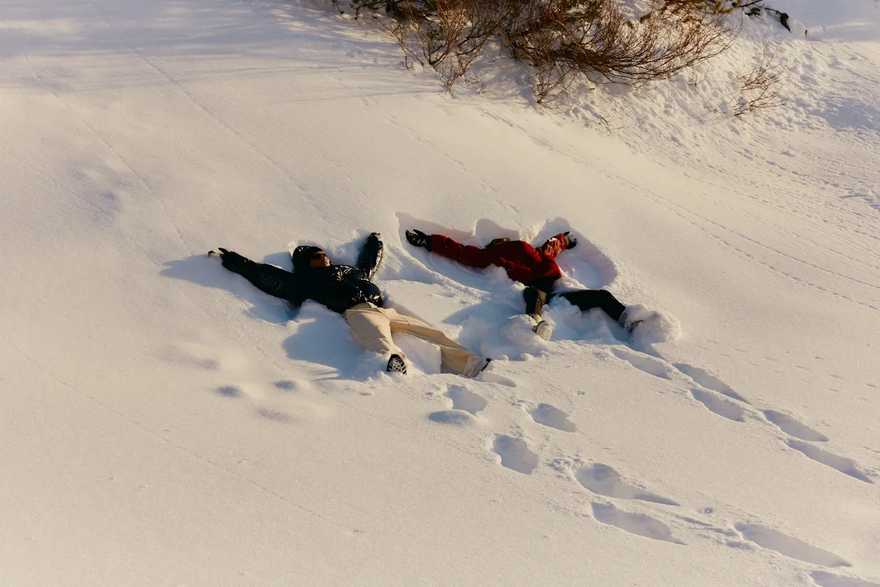 Two people lying on snow making snow angels under sunlight near leafless bushes.