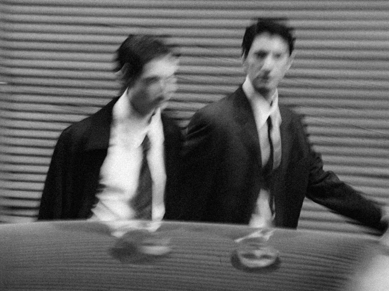 Black and white blurred photo of two men in suits walking beside a corrugated wall, reflected on a shiny surface.