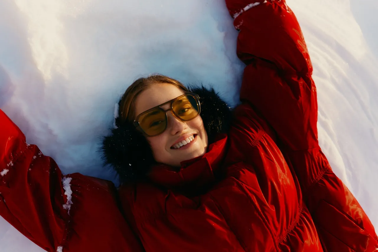 Smiling woman lying on snow wearing a red winter jacket, black earmuffs, and yellow-tinted sunglasses.