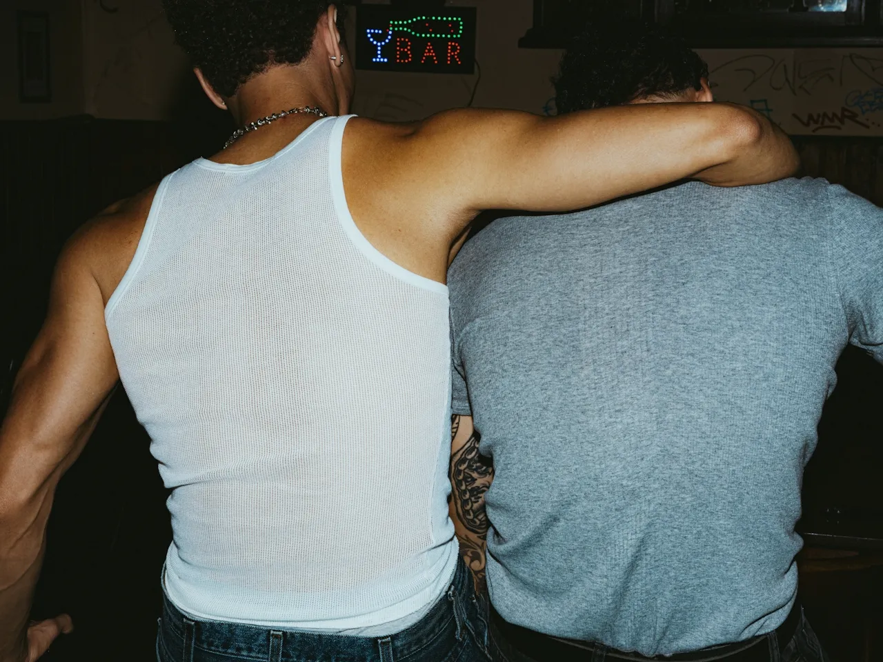 Two men seen from behind, one in a white tank top with his arm around the shoulder of the other man wearing a gray t-shirt, with a neon bar sign in the background.