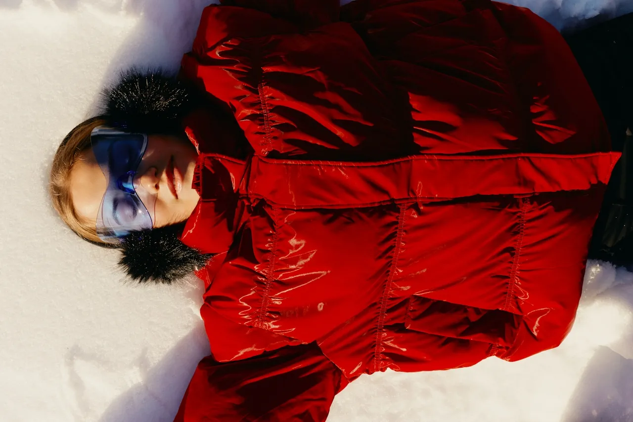 Person lying on snow wearing a red puffer jacket, blue ski goggles, and black fur earmuffs.