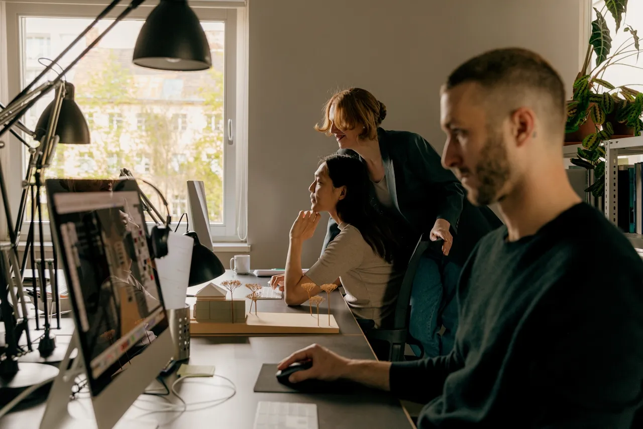 Three coworkers working at desks with computers in a bright office with large windows and plants.