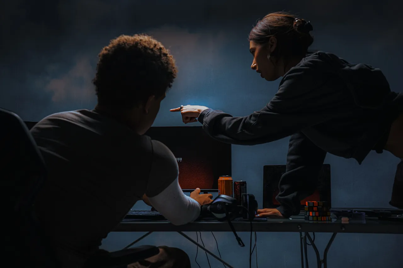 Two people focused on a computer screen in a dimly lit room, with one pointing at the monitor and various items like headphones and a Rubik's cube on the desk.