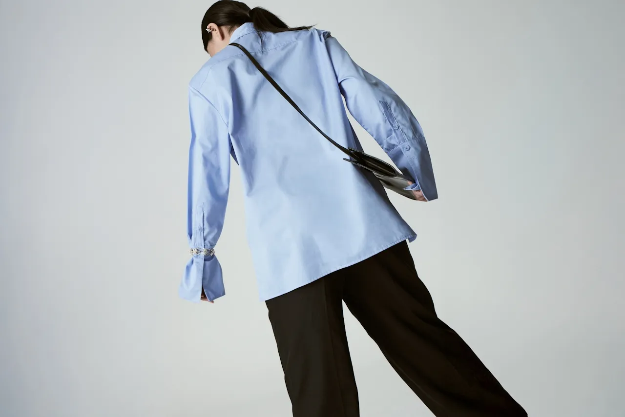 Person wearing a light blue long-sleeve shirt and black pants, carrying a small black shoulder bag, photographed from behind against a plain background.
