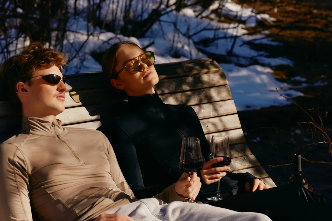 Man and woman wearing sunglasses relaxing on a wooden bench outdoors with snow and holding glasses of red wine.
