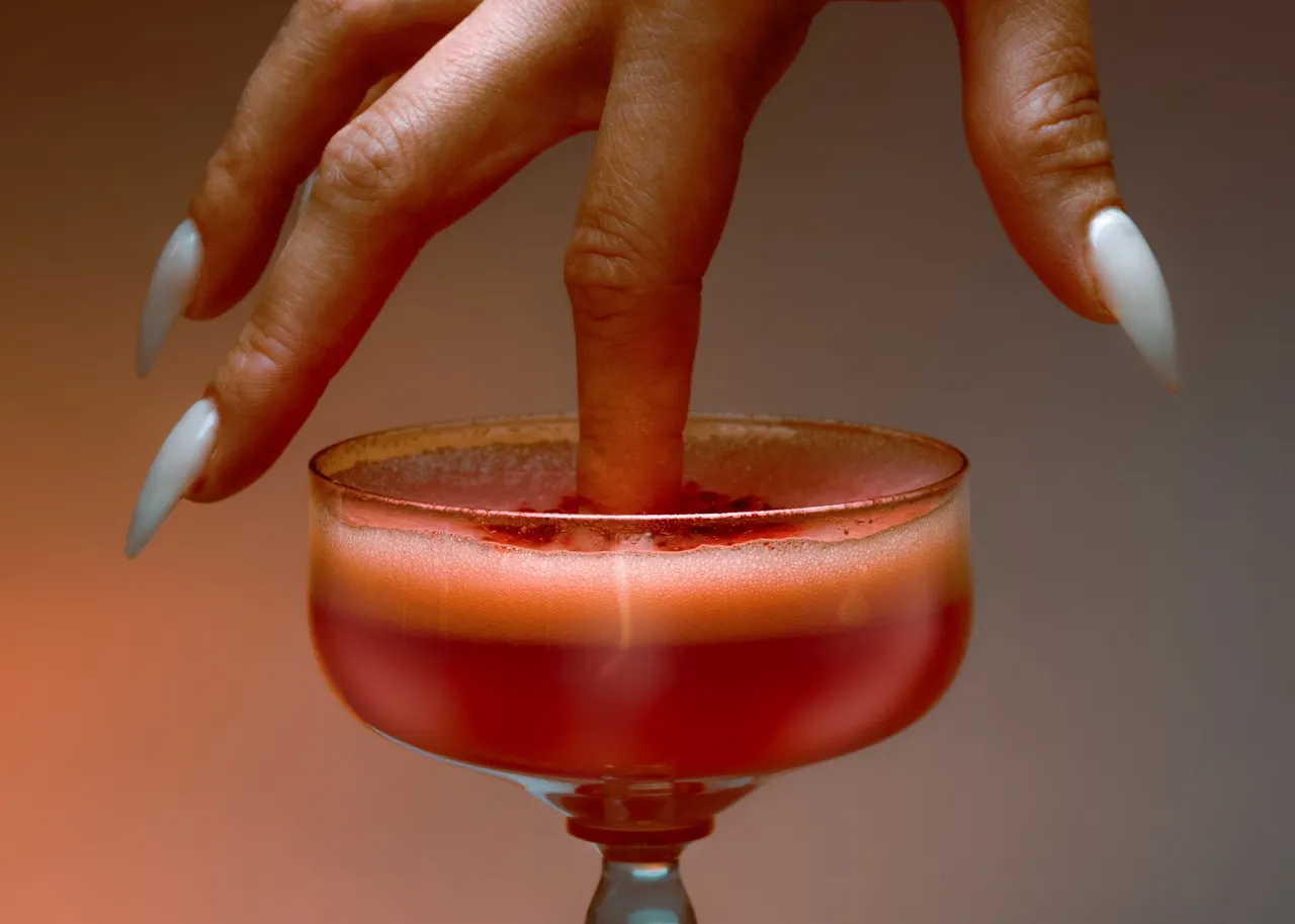 Hand with long white nails dipping a finger into a red frothy cocktail in a coupe glass.