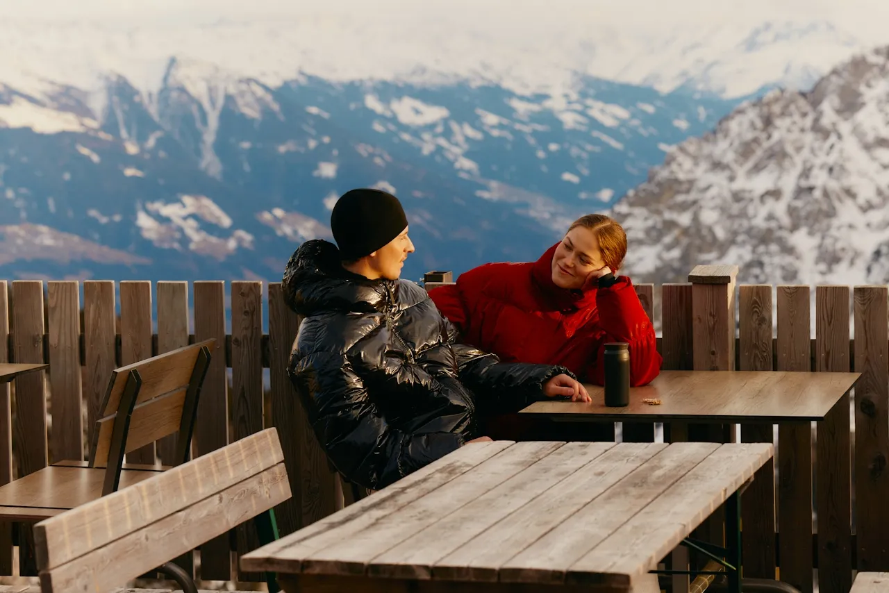 Two people in winter jackets sitting and talking at a wooden table outdoors with snow-covered mountains in the background.