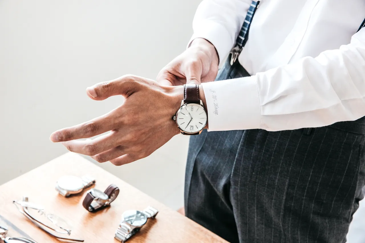 Person adjusting a wristwatch with a brown leather strap, standing near a wooden table displaying multiple watches and eyeglasses.