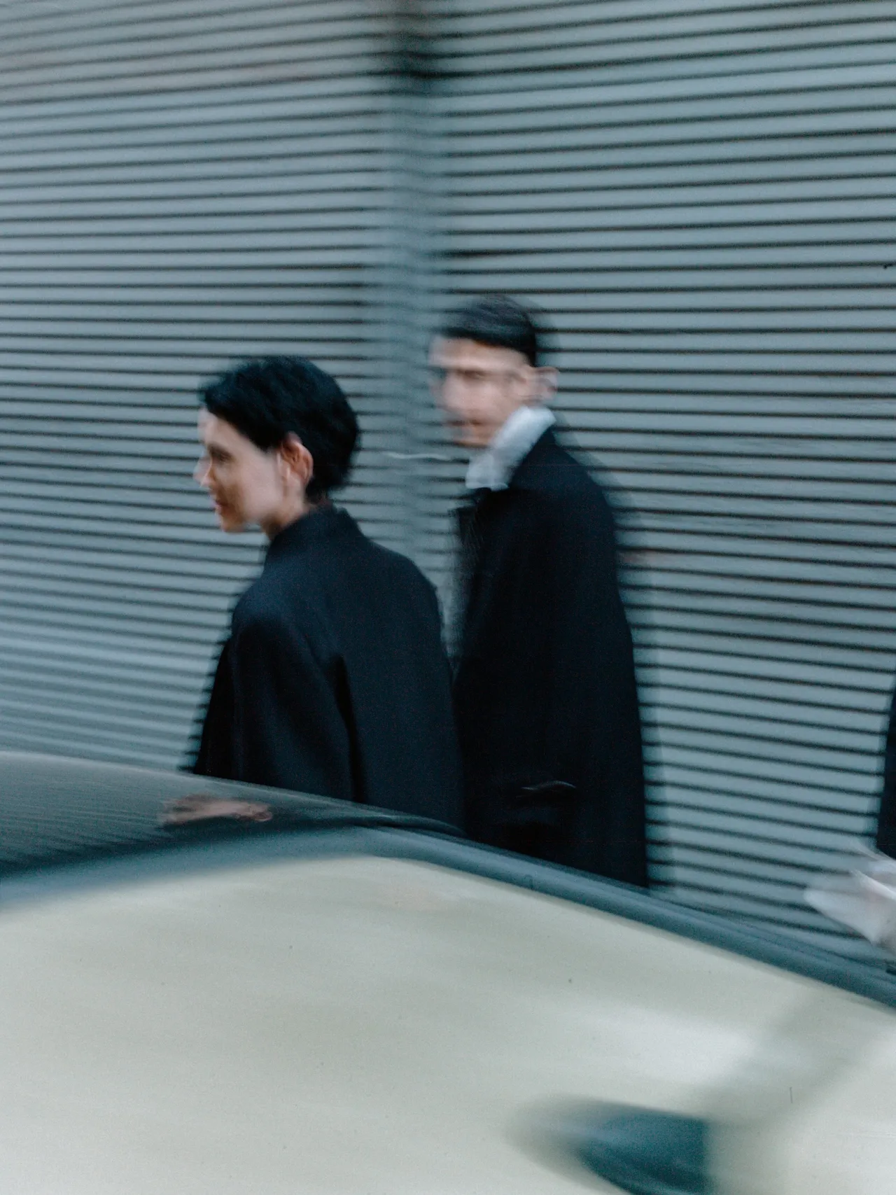 Blurry photo of two people walking past a grey corrugated metal wall with a car roof in the foreground.