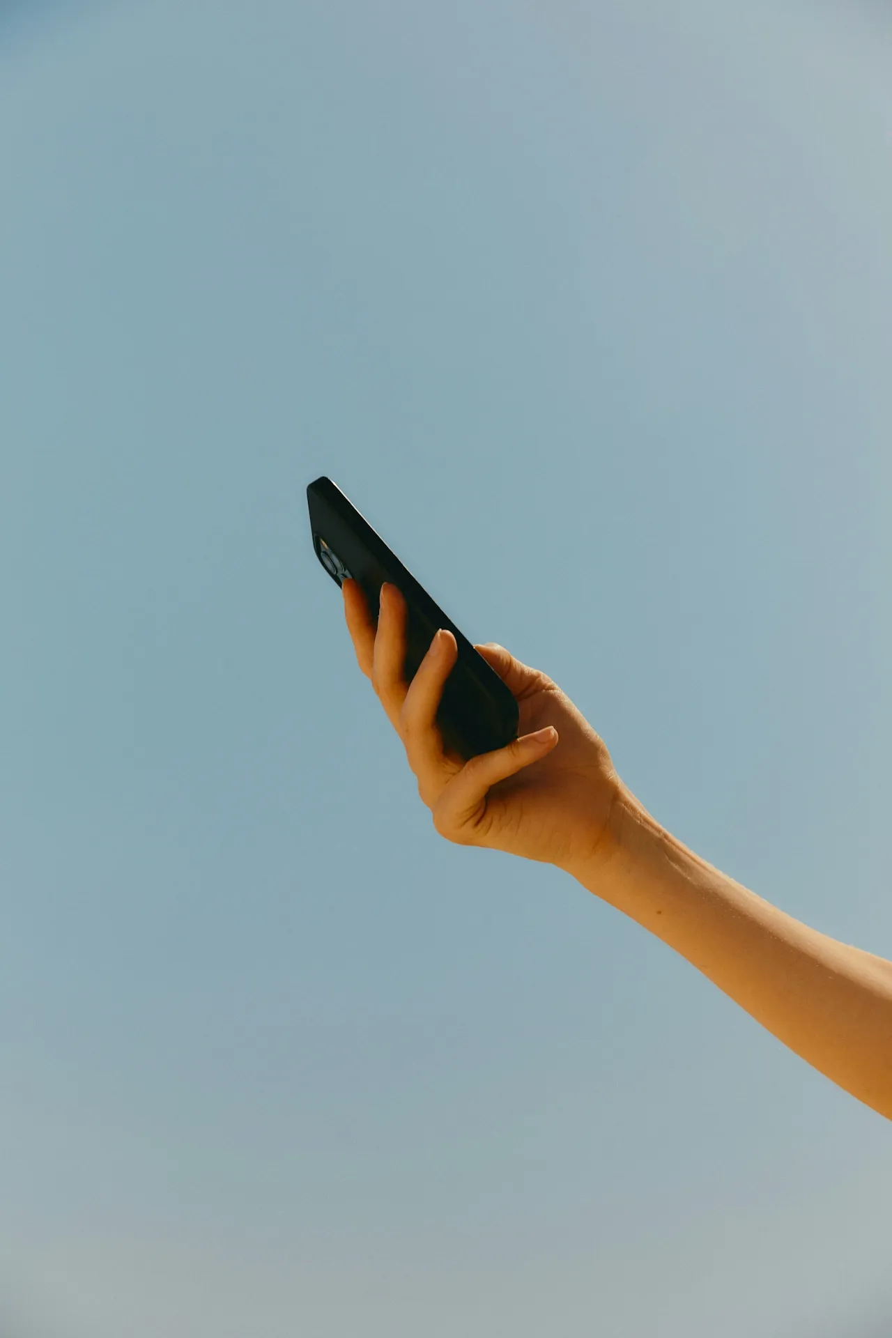 Hand holding a black smartphone against a clear blue sky.