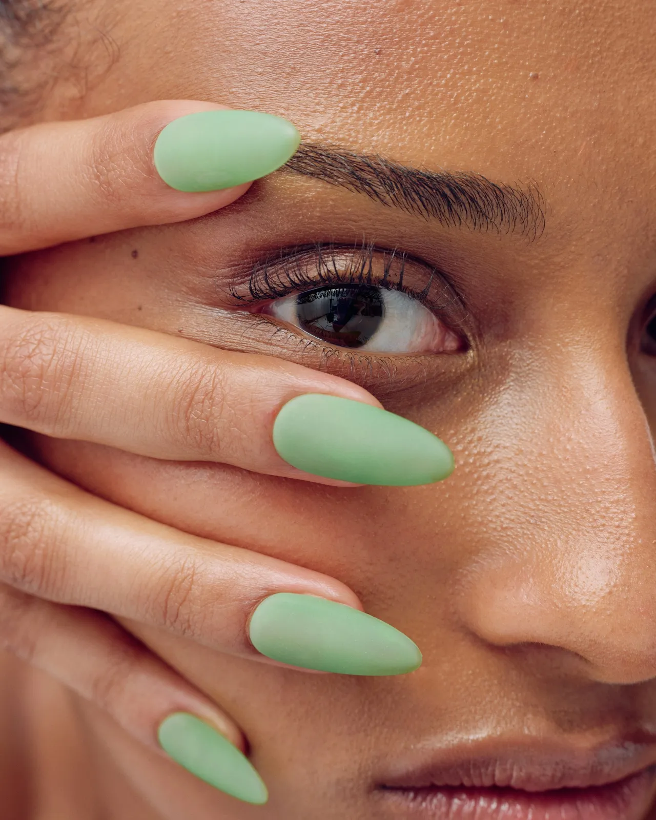 Close-up of a face with a hand showing long matte green almond-shaped nails near the eye and cheek.