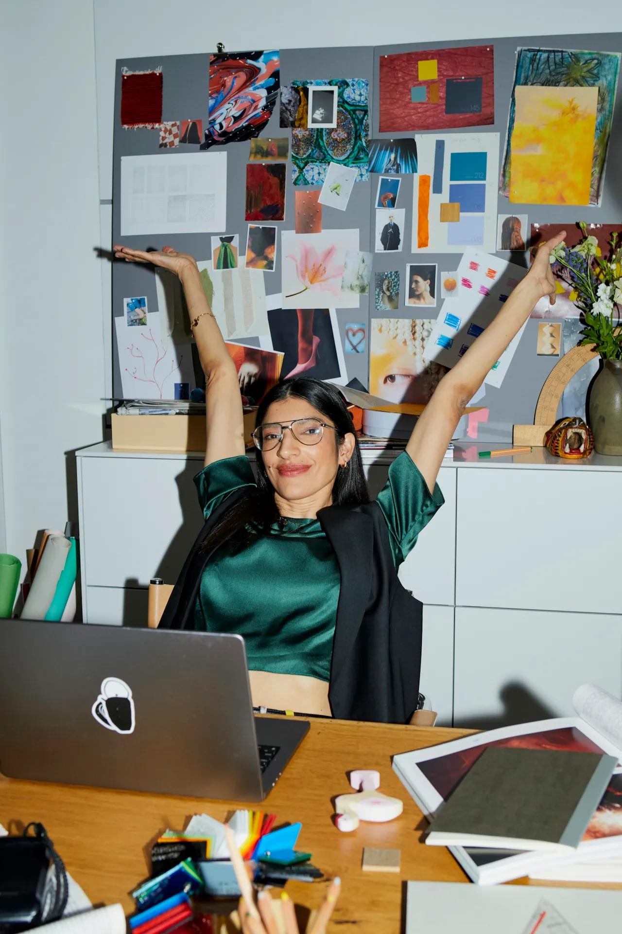 Smiling woman with glasses sitting at a desk with laptop and art supplies, stretching arms upward in front of a collage-covered board.
