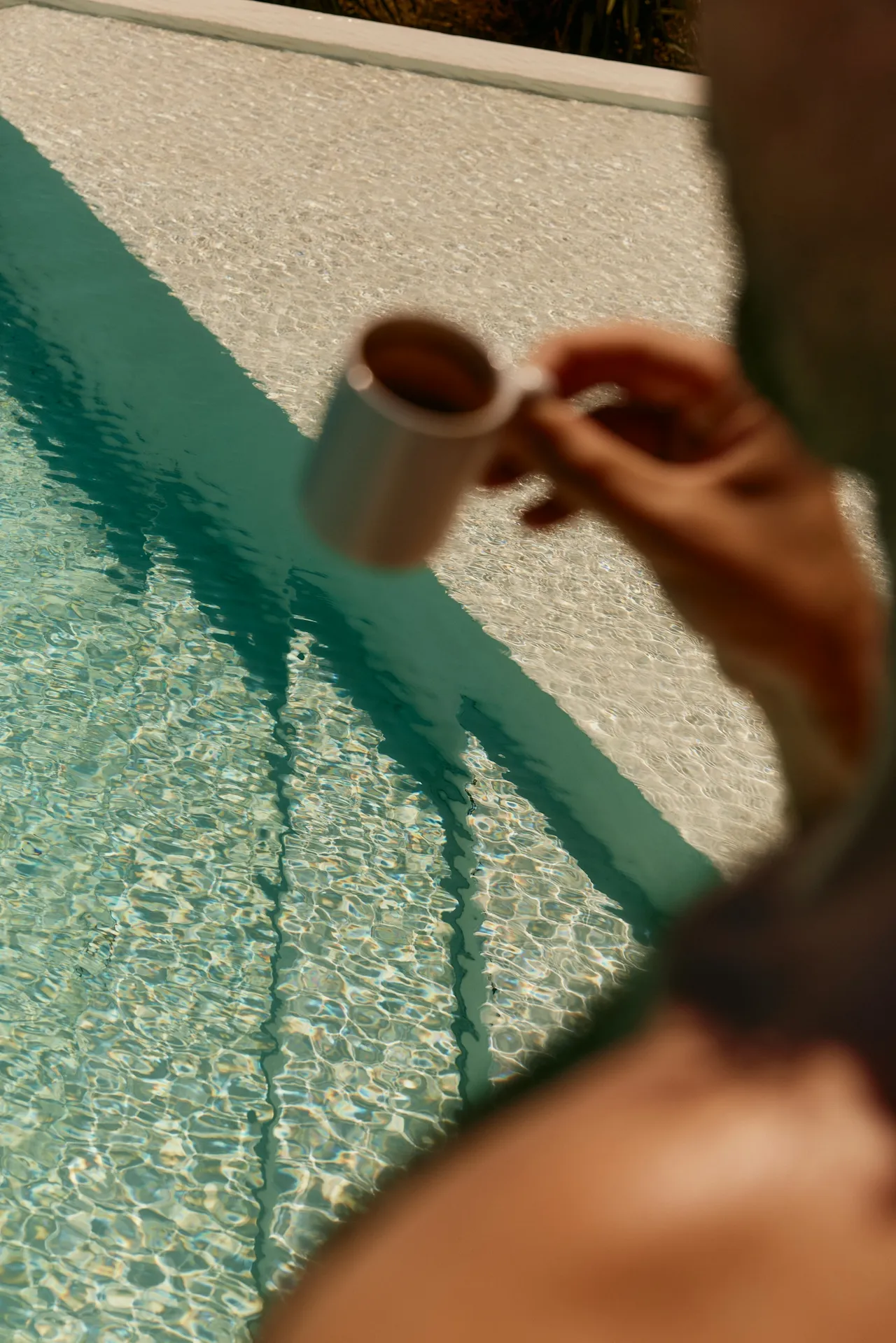 Person holding a small white cup by a sunlit swimming pool with clear blue water and tiled steps.