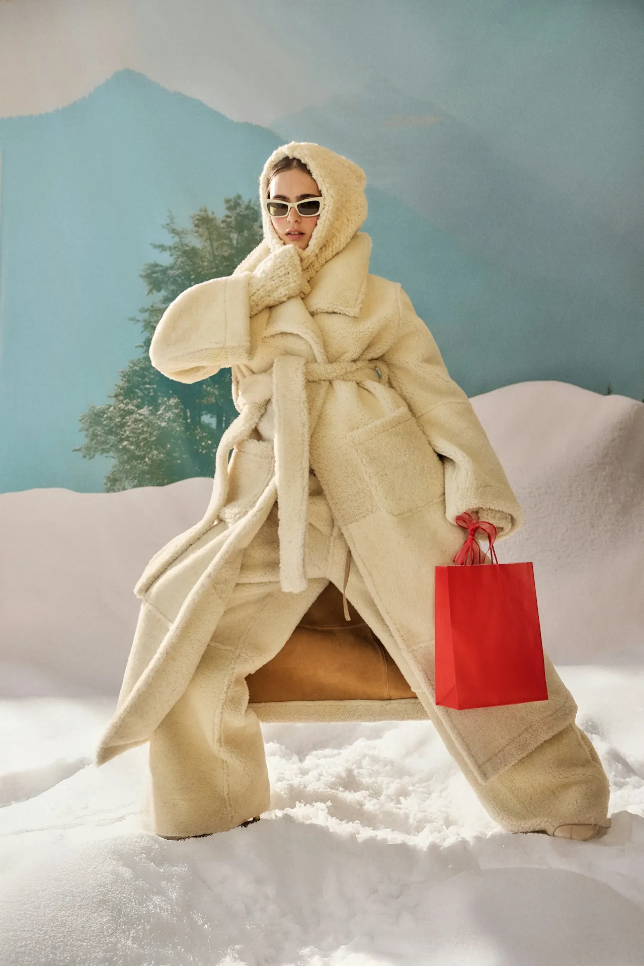Woman in a cream-colored hooded shearling coat and matching pants standing on snow, holding a red shopping bag.