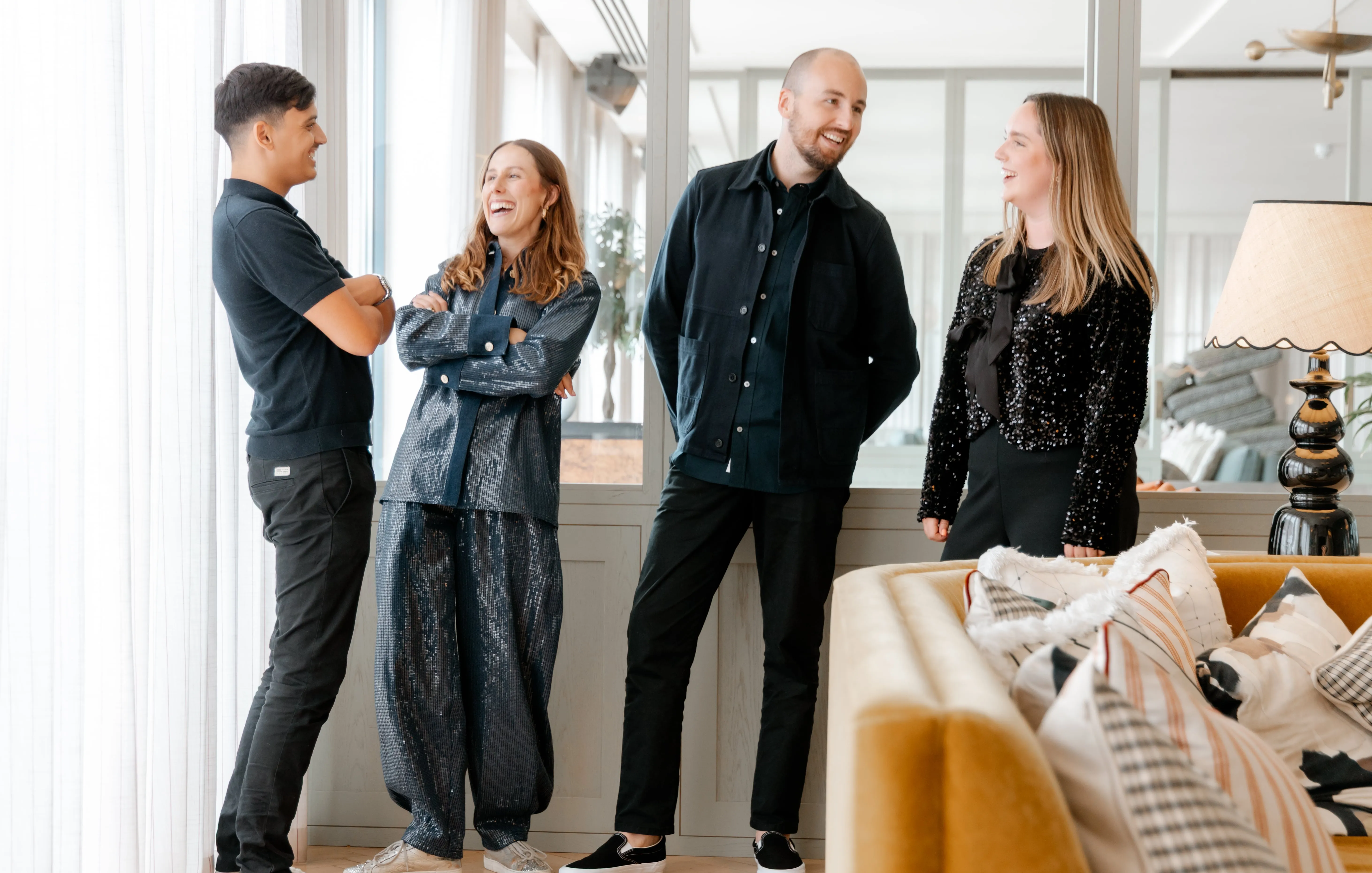 Four young adults standing indoors near a window, smiling and engaged in conversation in a modern, well-lit room.