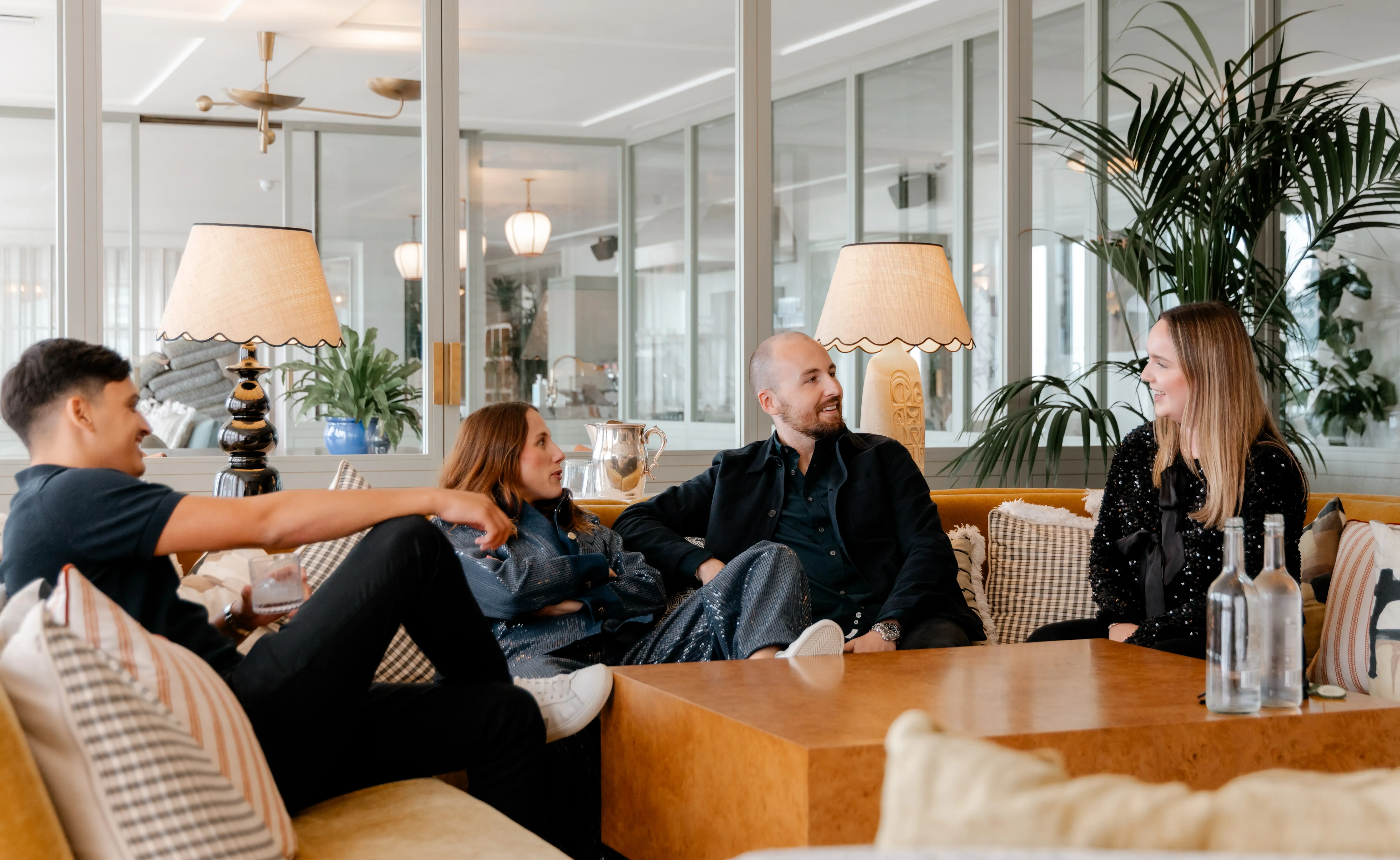 Four people sitting on couches in a modern, well-lit lounge area, engaged in conversation with two table lamps and plants in the background.