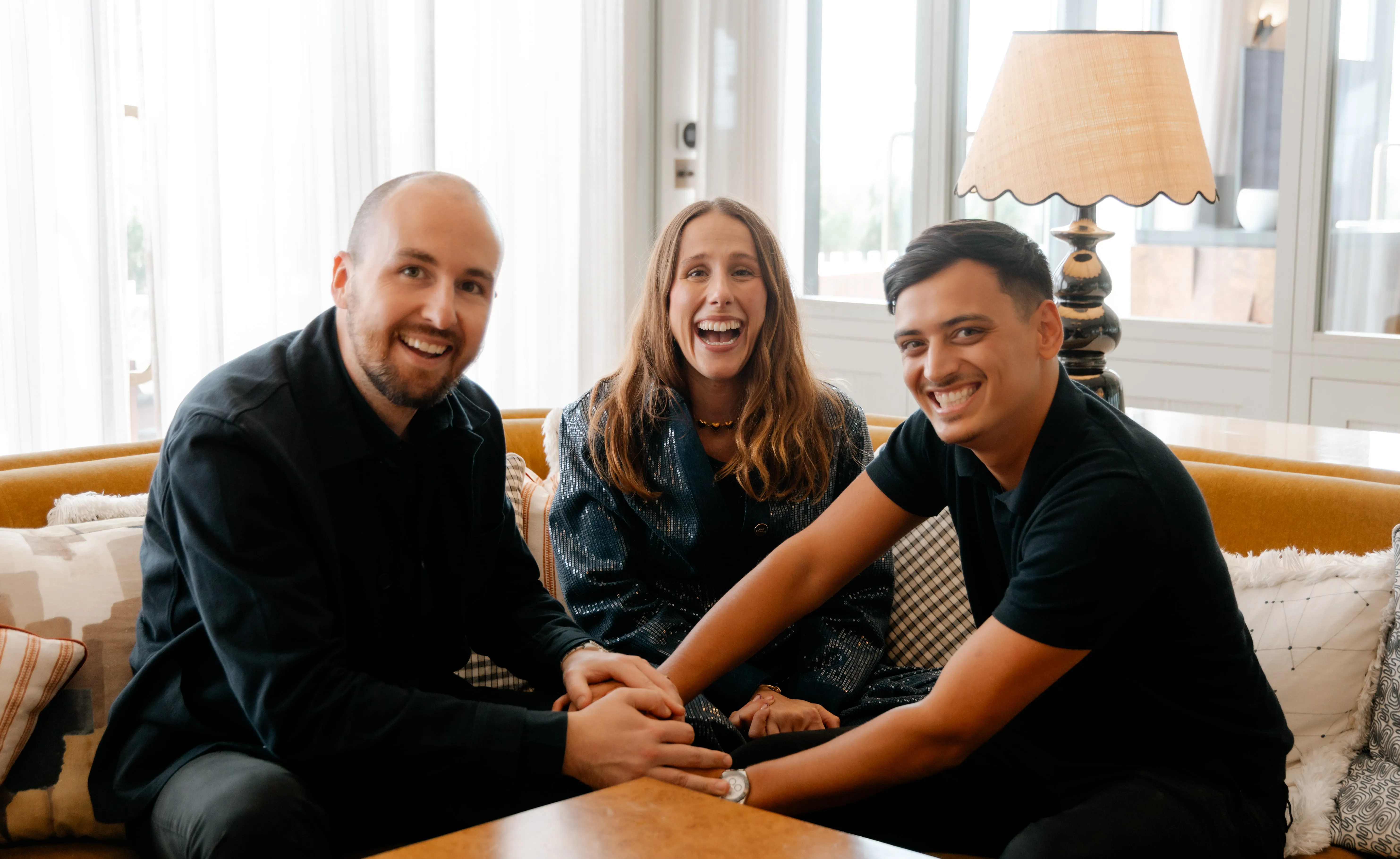 Three smiling adults sitting on a couch indoors with their hands stacked together on a table.