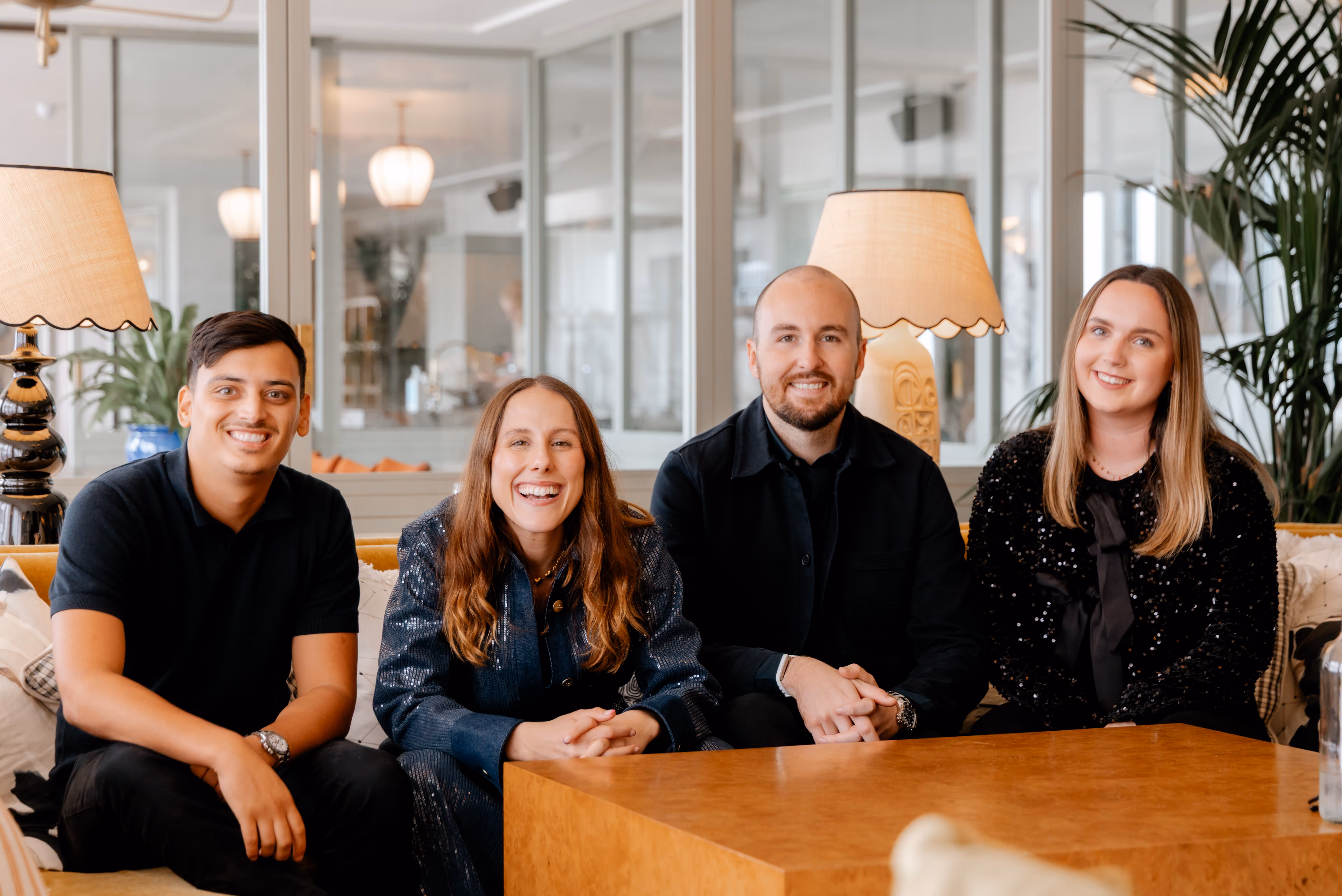 Four smiling people seated on a couch in a modern, well-lit office with lamps and plants in the background.