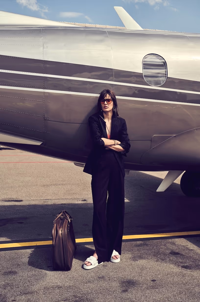 Woman in black outfit leaning against private jet with travel bag.