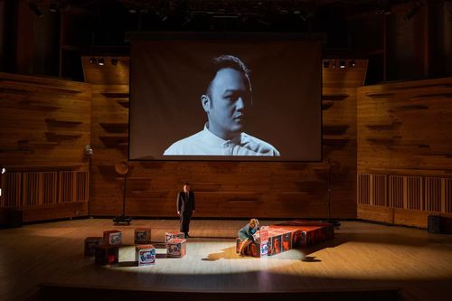 A stage with wooden walls featuring two people, a man standing and a woman crouching next to boxes decorated with Brillo and Kellogg's Corn Flakes designs, under a large screen projecting a black and white portrait of a man in a white shirt.