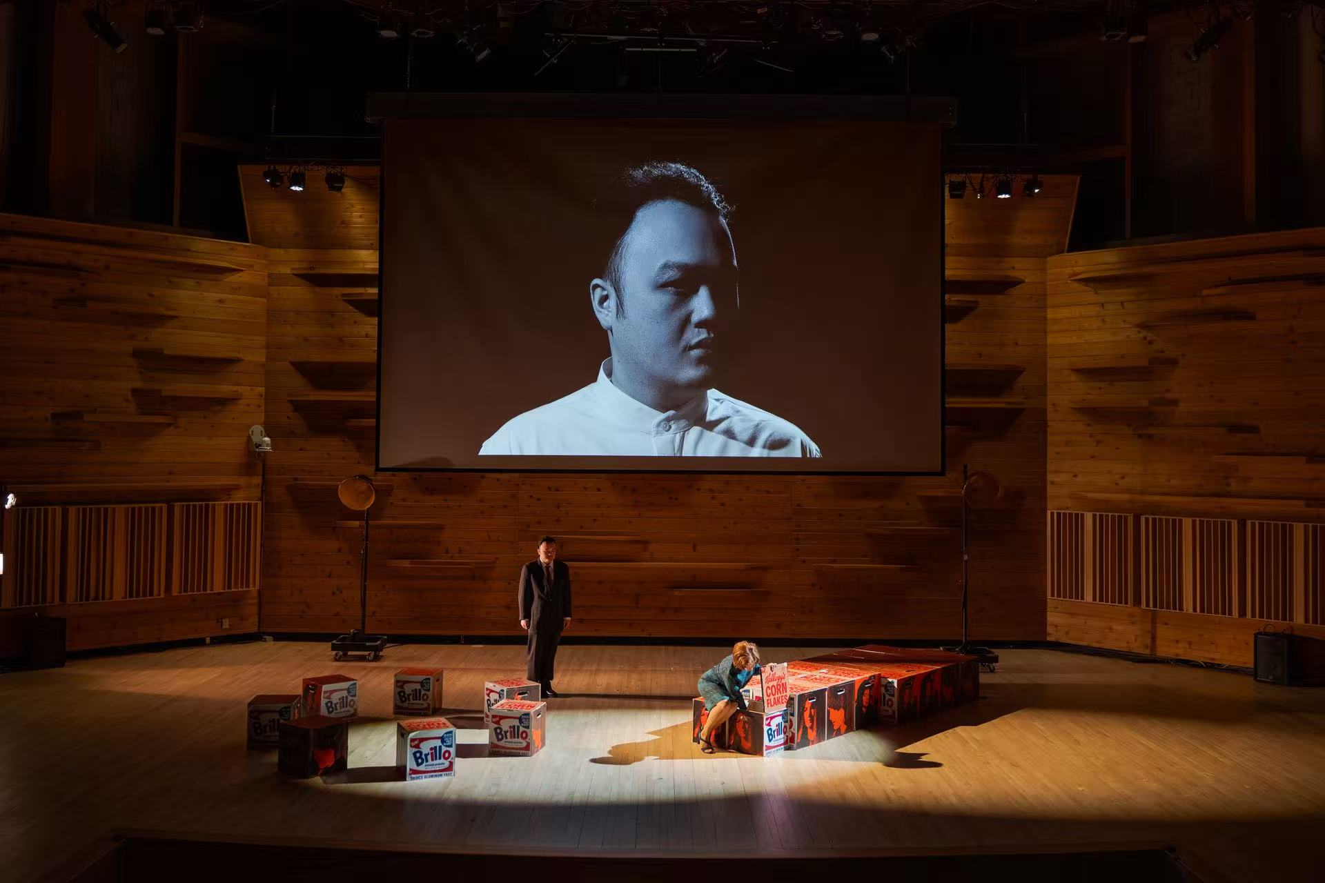 A stage with wooden walls featuring two people, a man standing and a woman crouching next to boxes decorated with Brillo and Kellogg's Corn Flakes designs, under a large screen projecting a black and white portrait of a man in a white shirt.
