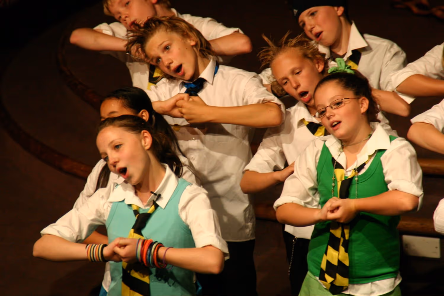 Group of children in school uniforms performing a song and hand gesture on stage.