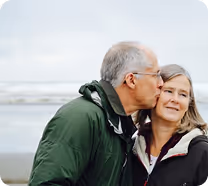 Older man in green jacket kissing woman with long hair on the cheek by the sea.
