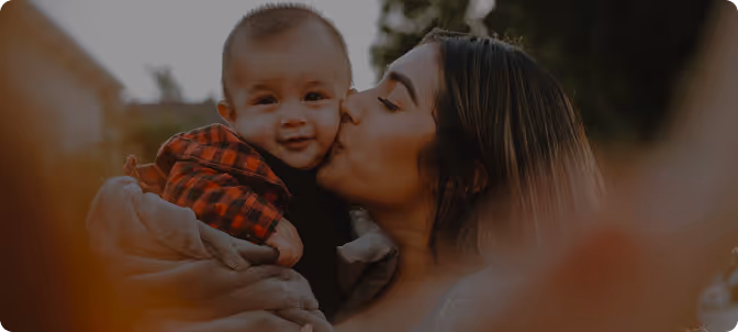 Woman holding and kissing a smiling baby wrapped in a blanket outdoors.