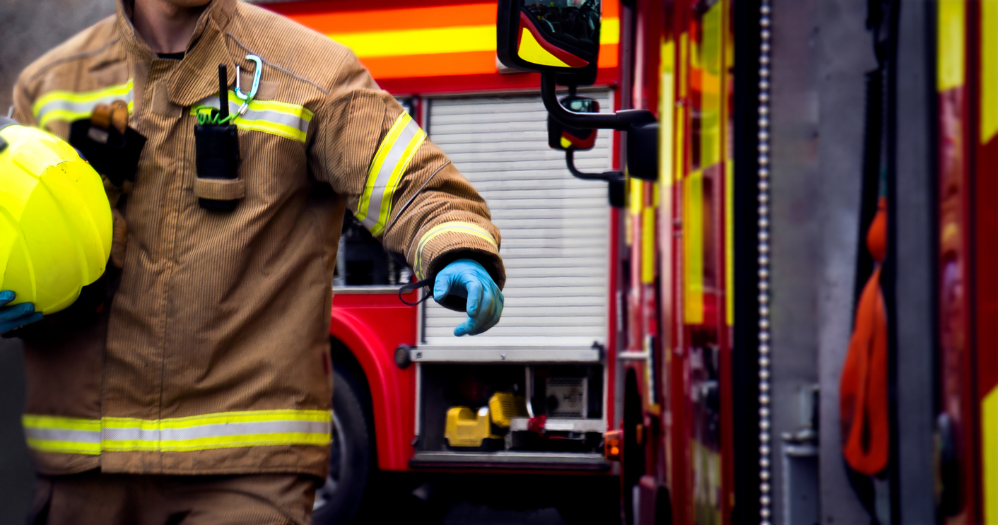 Firefighter in beige turnout gear holding a yellow helmet next to a red fire truck.